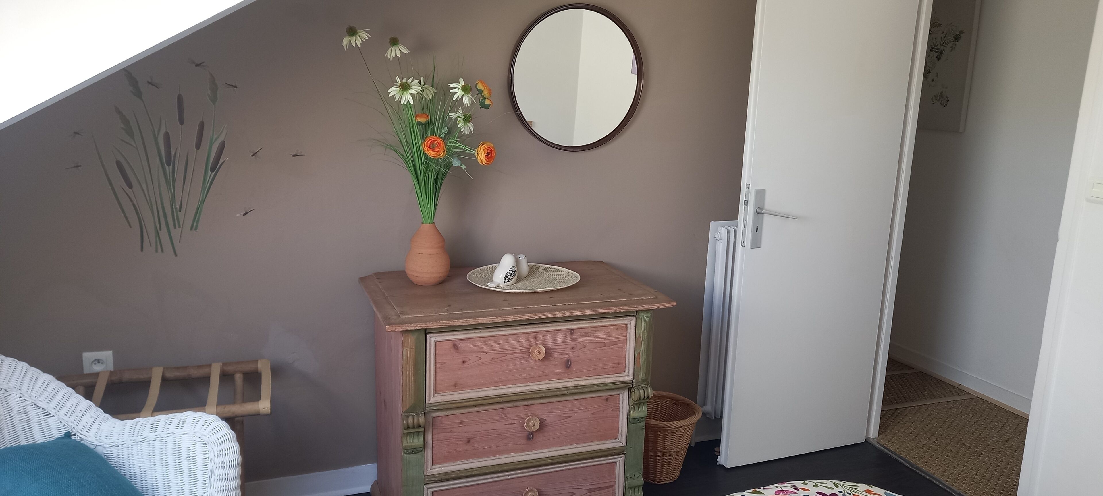 A bedroom with a dresser, a mirror, a vase with flowers, and a bed.