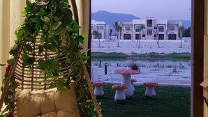 A room with a hanging chair, a wooden table, and a view of a building and landscape through a large window. - BAMBOO HAWANA Salalah - Forest Island  (Salalah)