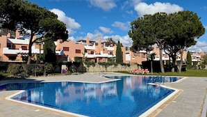 A swimming pool with lounge chairs, surrounded by trees and buildings with a clear blue sky above. - Spacious 1-Bedroom Apartment with Private Rooftop & Sunny Terrace (Quarteira)