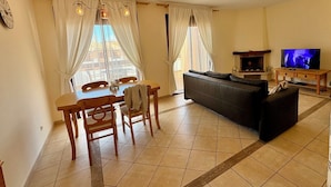 A living room with a sofa, a dining table with chairs, a TV, and a fireplace. - Spacious 1-Bedroom Apartment with Private Rooftop & Sunny Terrace (Quarteira)