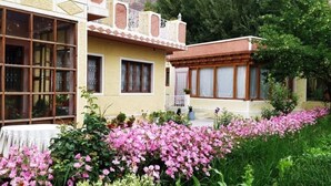 A two-story building with large windows and a balcony, surrounded by a garden with pink flowers. - Trinity Home Stay (Leh)