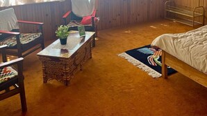 A cozy room with a wooden ceiling, a bed, a window with sheer curtains, a small table with a plant, and a chair. - Trinity Home Stay (Leh)