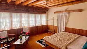 A cozy room with a wooden ceiling, a bed, a desk, a chair, and a window with curtains. - Trinity Home Stay (Leh)