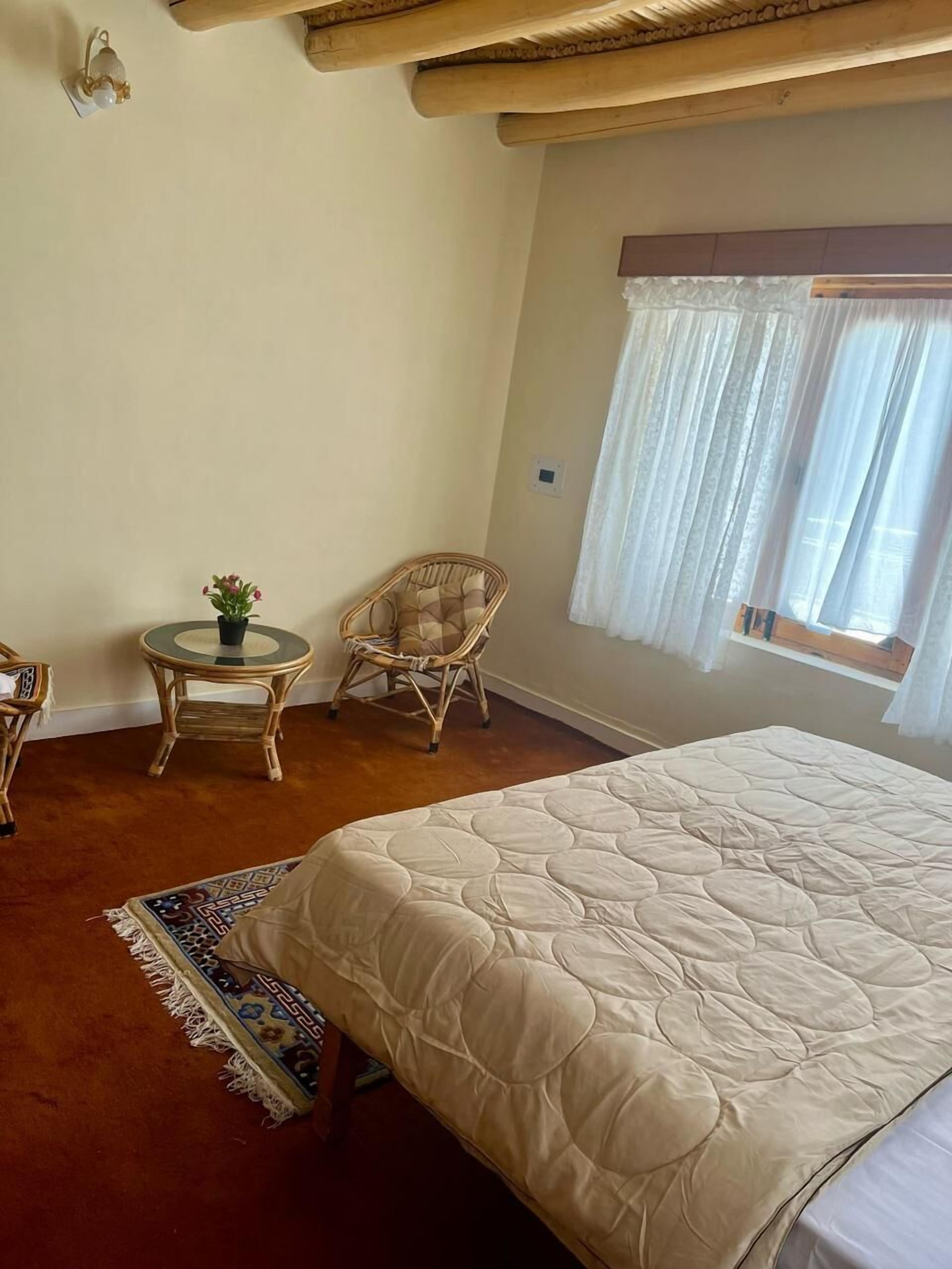 A bedroom with a large bed, a small round table with a plant, a wicker chair, and a window with sheer curtains.