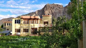 A two-story building with a flat roof, surrounded by lush greenery and mountains in the background. - Trinity Home Stay (Leh)