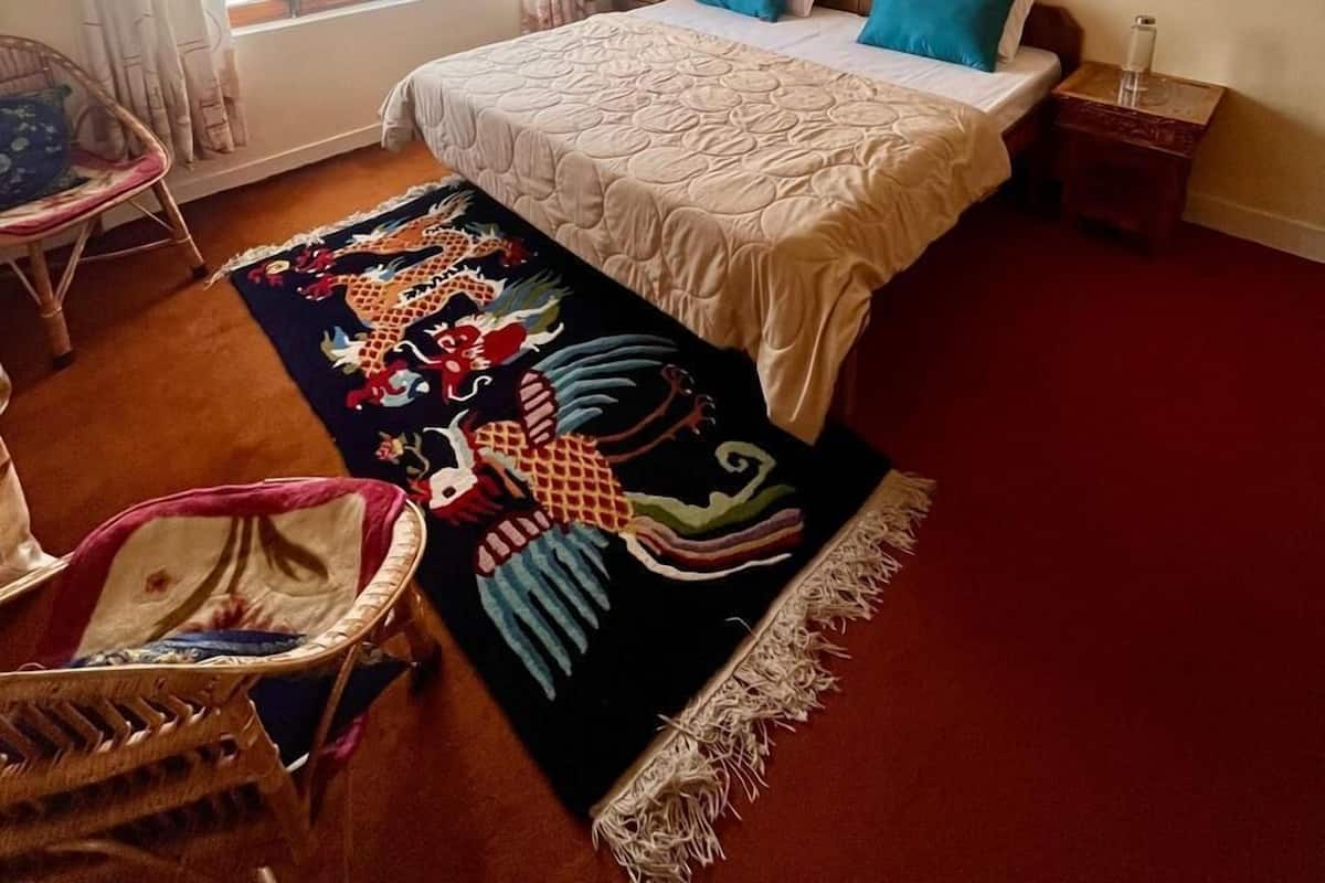 Un dormitorio con cama, dos almohadas, una mesita de noche, una silla, un ventanal con cortinas y una alfombra decorativa.
