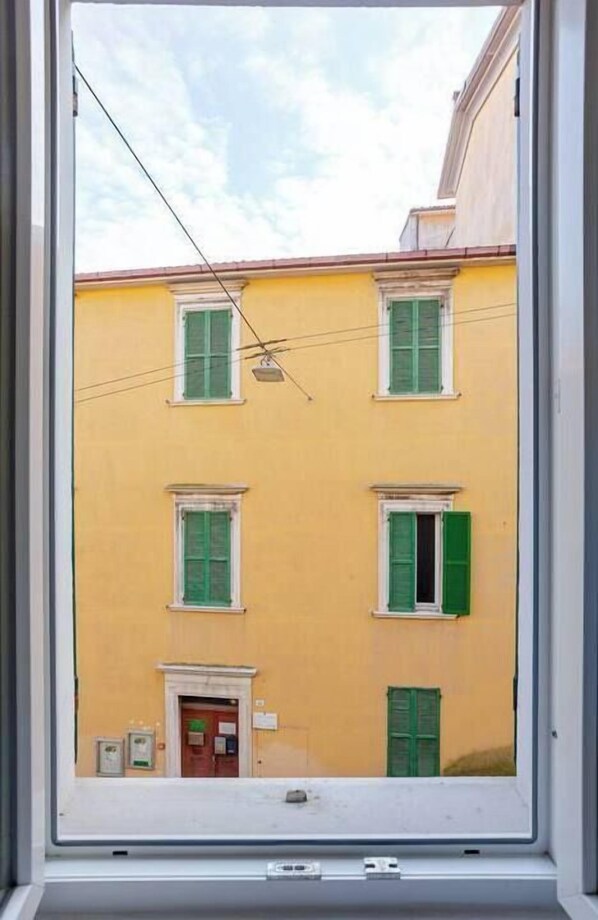 A window view of a yellow building with green shutters and a door, framed by a window with a white frame. - DA.NI.TO. ROOMS (Ancona)
