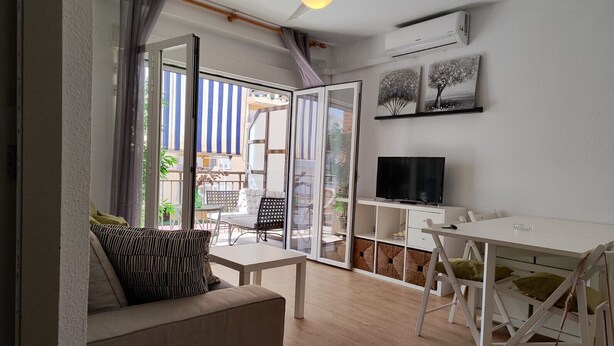A modern living room with a sofa, a dining table, a TV, and a balcony with outdoor seating.