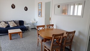 Ein Wohnzimmer mit einem dunkelblauen Sofa, einem kleinen weißen Tisch, einem Holzespeisetisch mit vier Stühlen und einer weißen Wand mit gerahmten Bildern und einem Spiegel.