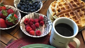 Un plato de gofres cubiertos de frutos rojos, un tazón de arándanos, un tazón de frambuesas y una taza de café sobre una mesa de madera.