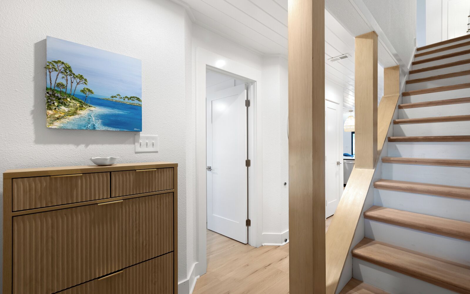 A hallway with a wooden staircase, a white door, and a painting of a beach scene.
