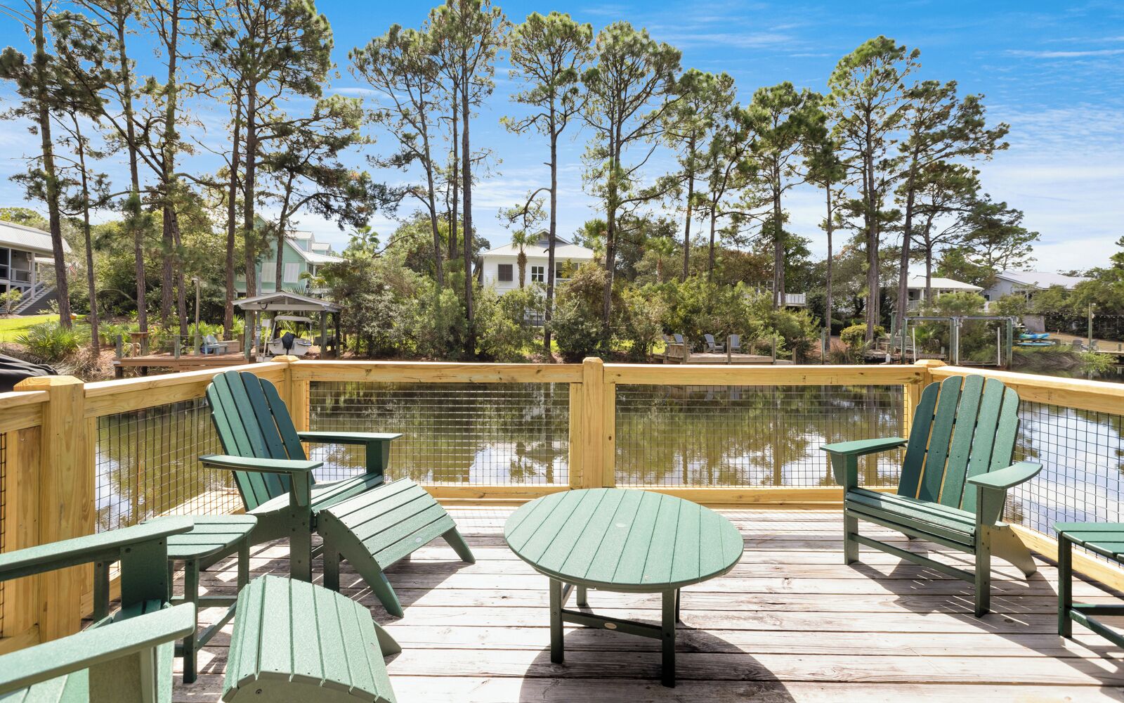 A wooden deck with green Adirondack chairs and a table, overlooking a calm body of water and surrounded by trees.