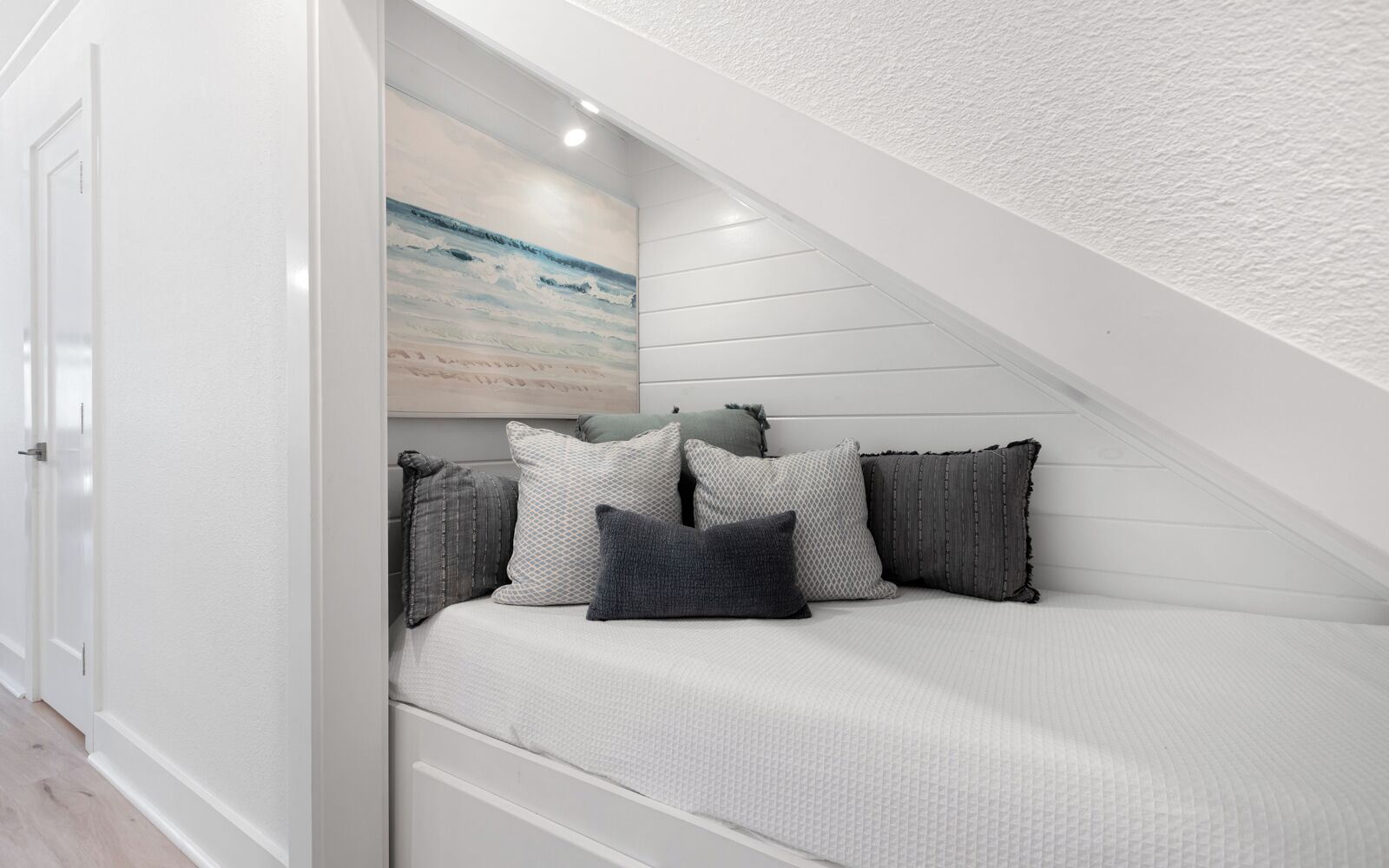 A white, sloped ceiling with a painting of a beach scene, a white bed with patterned bedding, and several pillows.
