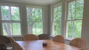 Ein Esszimmer mit Holztisch und Korbstühlen, großen Fenstern mit Blick auf Bäume und einer festen Bank an der Wand.