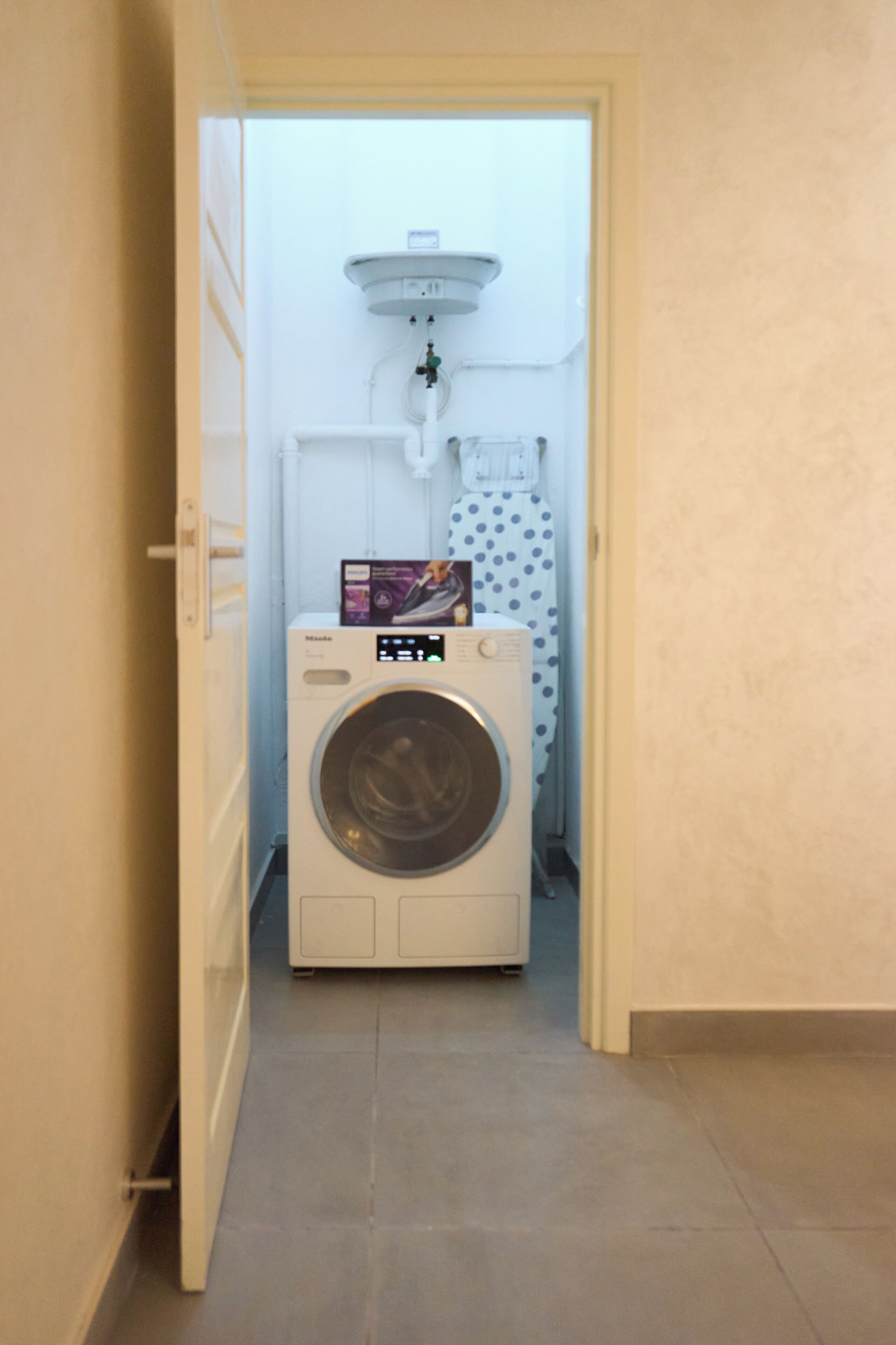 A laundry room with a front-loading washing machine, a small shelf with a box, and a towel rack with a towel.