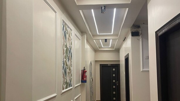 A hallway with a wooden floor, white walls, and a ceiling with linear lighting. There are doors on the right and a fire extinguisher on the left. - Hotel Lotus Inn (Varanasi)