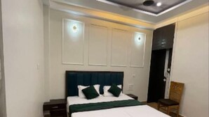 A modern bedroom with a wooden floor, a large bed with white bedding and green pillows, a ceiling fan, recessed lighting, and a dark-colored headboard. - Hotel Lotus Inn (Varanasi)