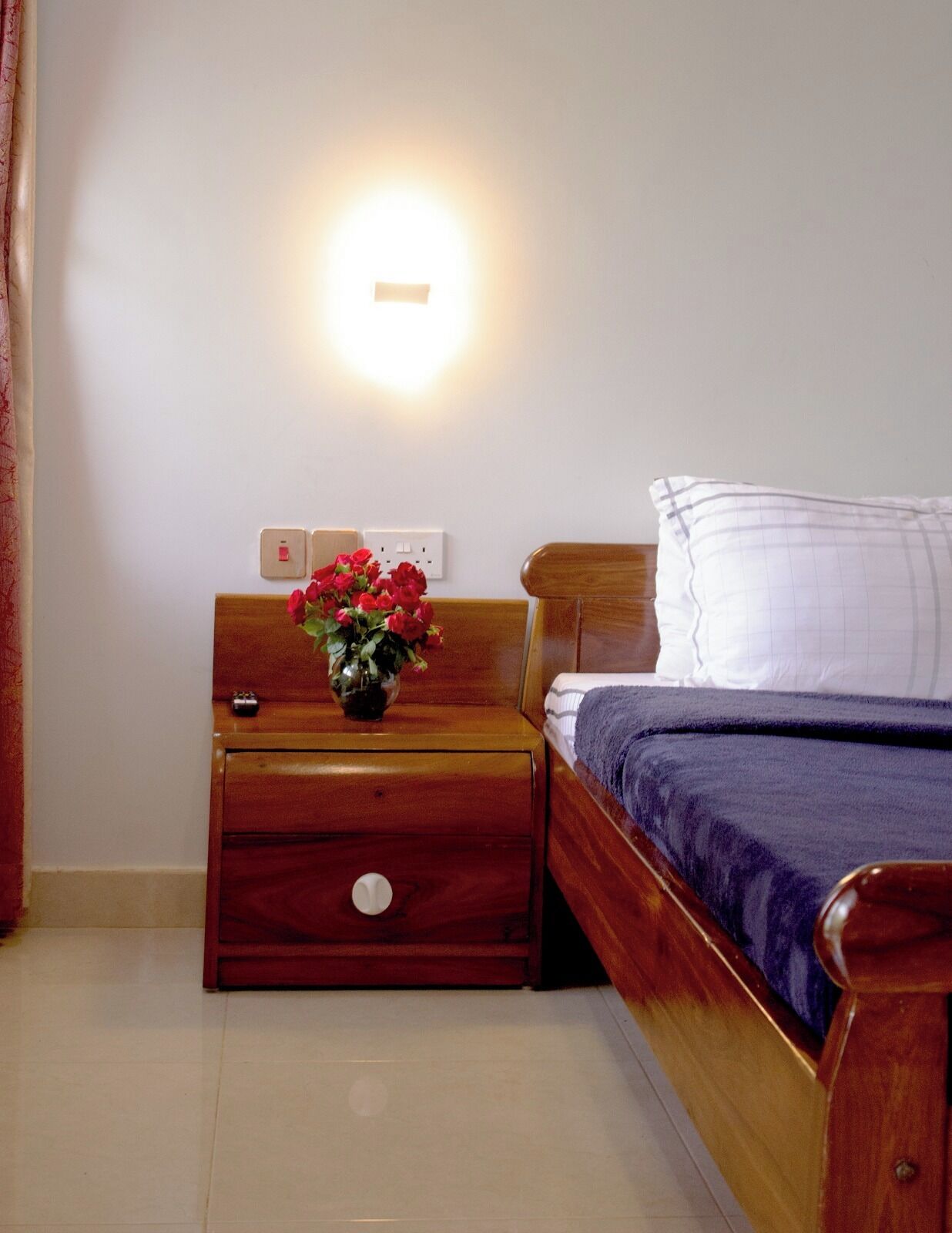 Un dormitorio con una mesita de noche de madera, una mesilla con un florero y una cama con una manta azul, además de una lámpara de pared.