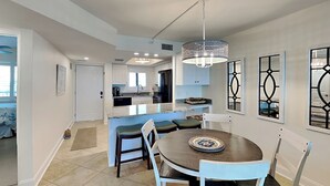 A modern kitchen with white cabinets, a central island, and a dining area with a round table and chairs. - Seaside 403 | New Gulf-Front Condo—Your Front-Row Seat to Paradise! (Fort Myers Beach)