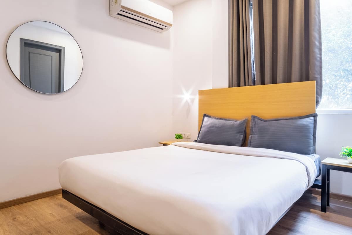 A neatly made bed with a wooden headboard, a round mirror on the wall, and an air conditioning unit above it.