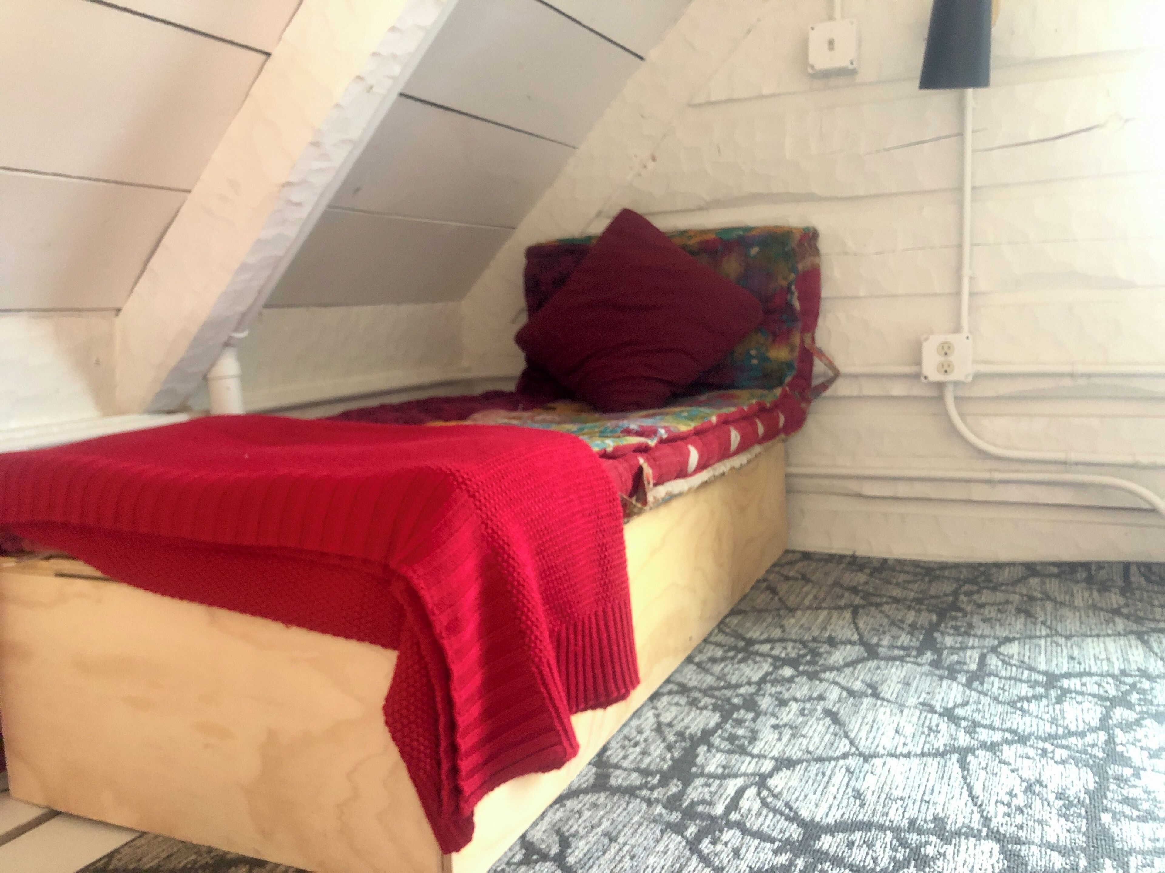 A small, cozy room with a sloped ceiling, a single bed with a red blanket, a colorful pillow, and a patterned rug.