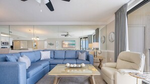 A living room with a sectional sofa, a glass coffee table, a beige armchair, a ceiling fan, and a view of the kitchen and dining area through a glass partition. - Phoenix X506  Feel the Sea Breeze (Orange Beach)