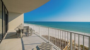 A balcony with a table and chairs, overlooking a beach with a clear view of the ocean and a pier in the distance.