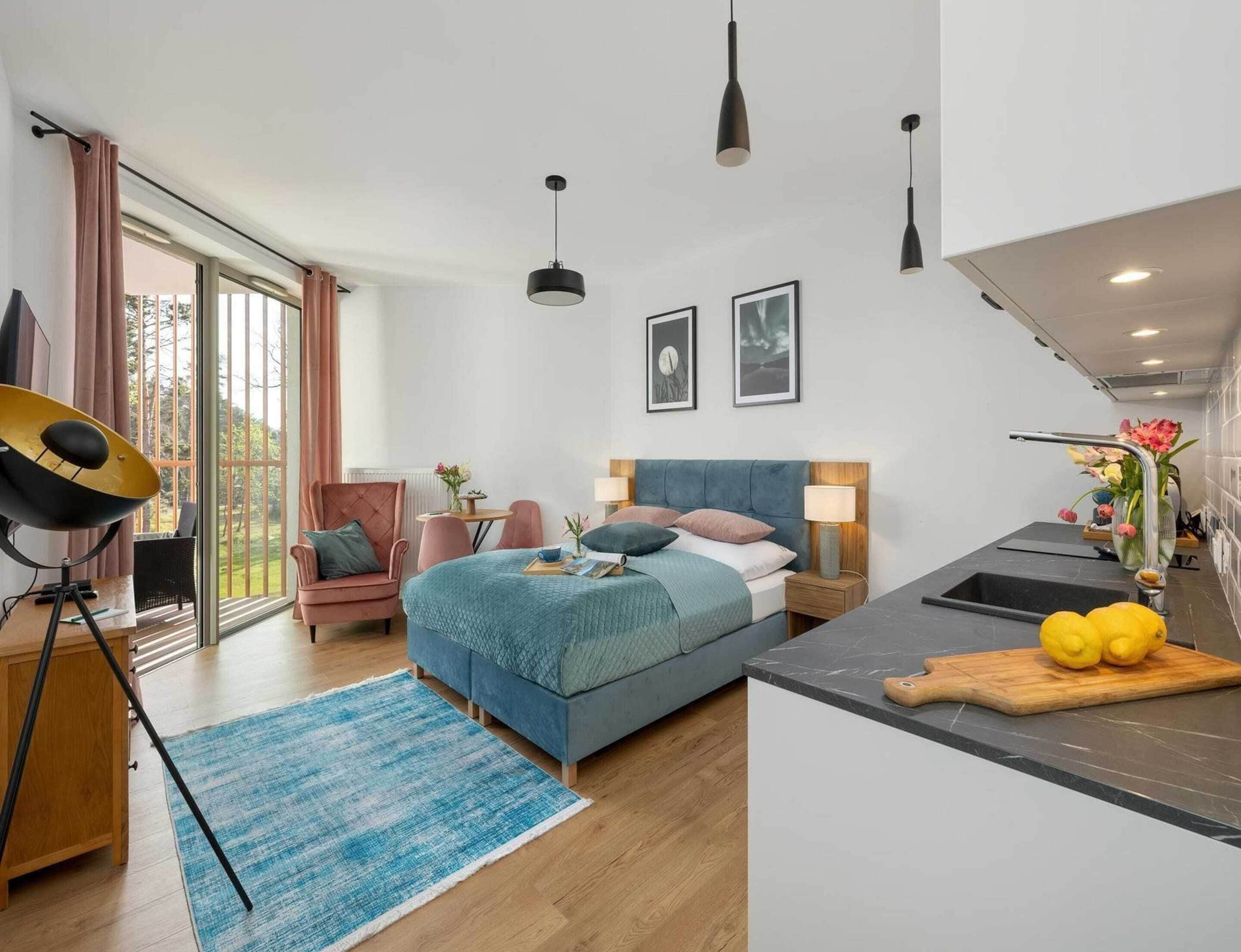 A modern hotel room with a bed, a desk, a chair, a dining table, and a kitchen area.