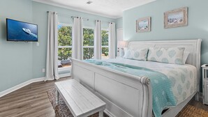 A bedroom with a bed, a bench, a ceiling fan, a TV, and a window with curtains.