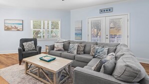 A living room with a grey sectional sofa, a wooden coffee table, a grey armchair, and a ceiling fan. There are two windows with blinds, a door with a cityscape view, and a wall-mounted star decoration.