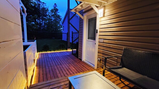 Una terraza de madera con una banca y una mesa, una puerta y un pequeño porche iluminado con luces de cuerda.