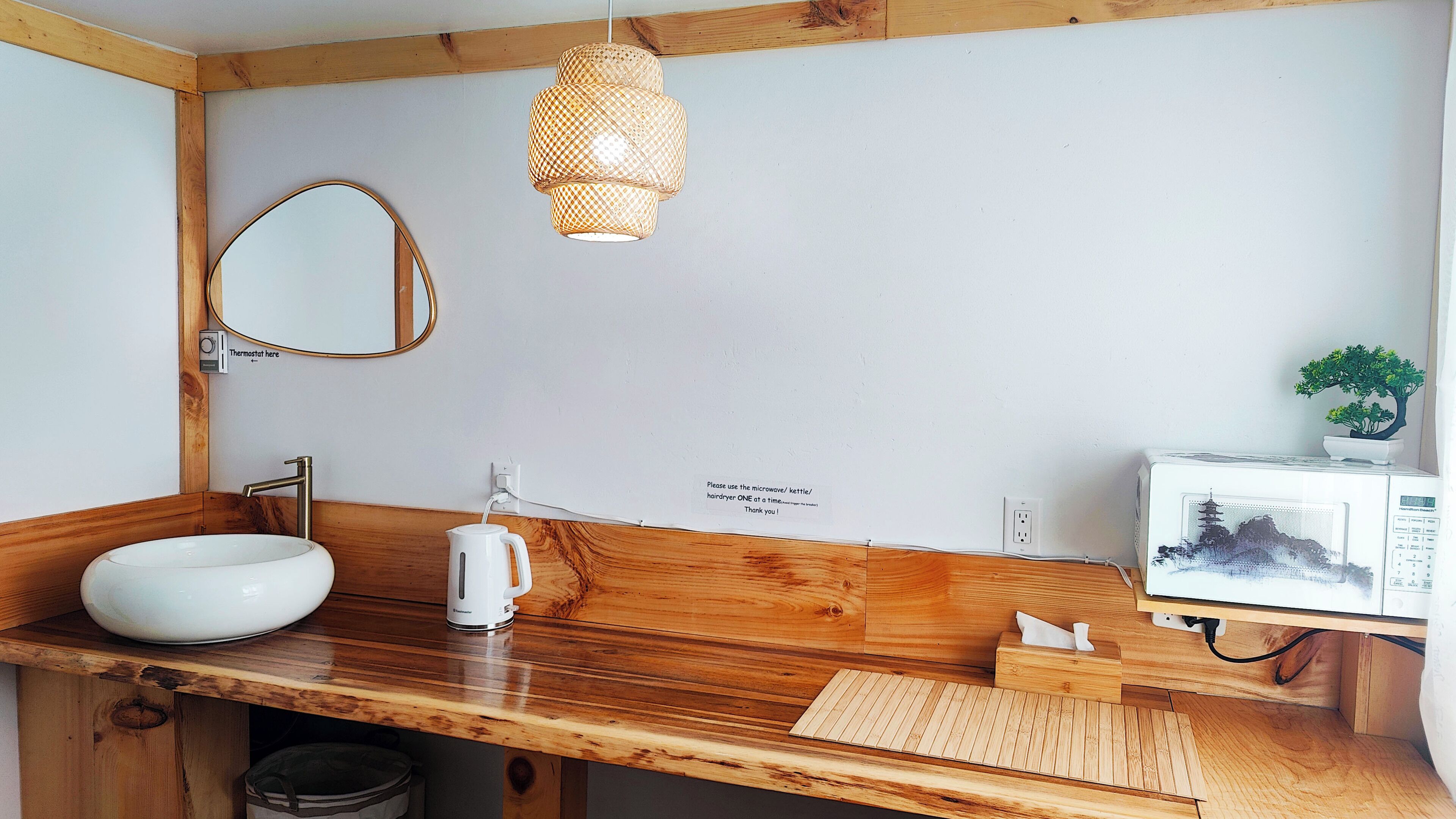 Une salle de bain avec un meuble-lavabo en bois, un lavabo blanc, un miroir, une bouilloire, un four à micro-ondes, une petite plante en pot et un panneau indiquant qu’il faut utiliser le four à micro-ondes, la bouilloire ou le sèche-cheveux un à la fois.