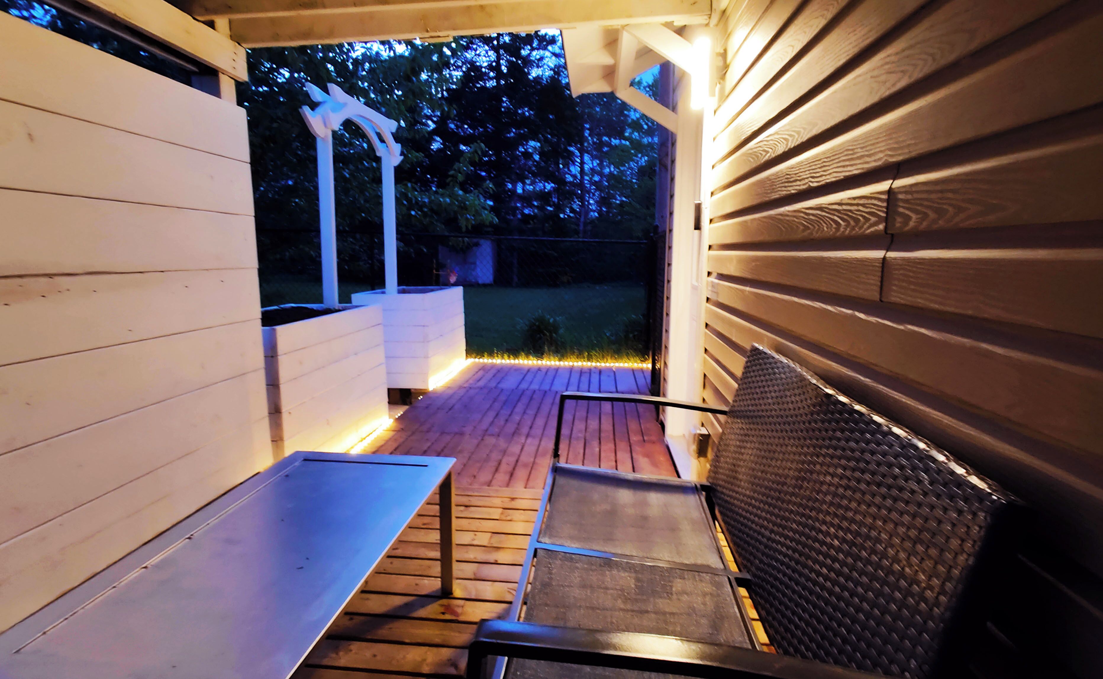 Un área de patio al aire libre con cubierta de madera, una mesa metálica y un banco de fibras trenzadas.