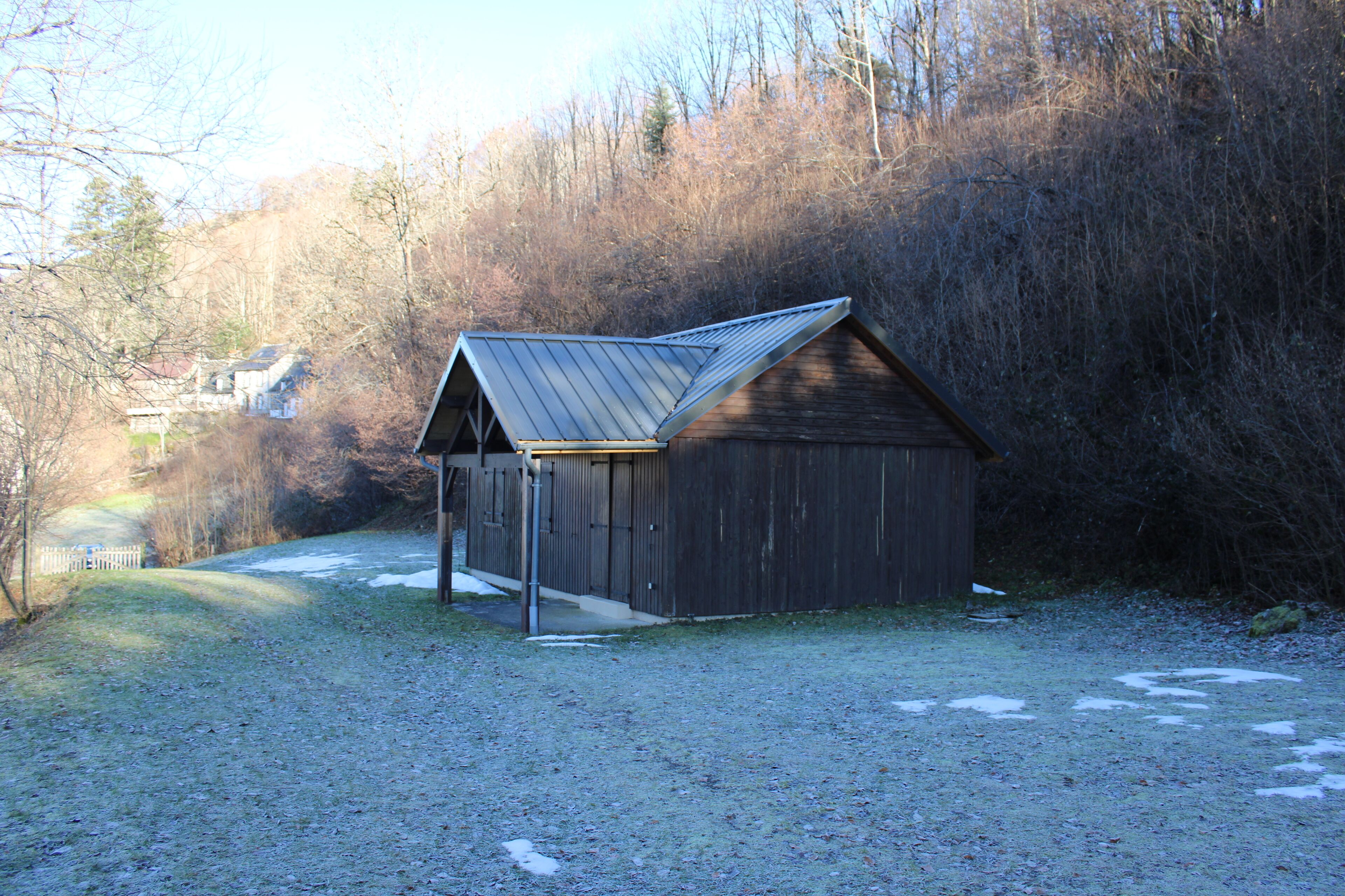 A wooden shed with a sloped roof, located in a grassy area with a path leading to a house in the background.