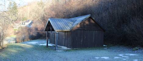 A wooden shed with a sloped roof, located in a grassy area with a path leading to a house in the background.