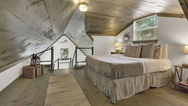Ein Schlafzimmer mit schrägverdeckter Decke, einem großen Bett, einem Fenster mit Jalousien und einer Treppe, die zu einer oberen Ebene führt.