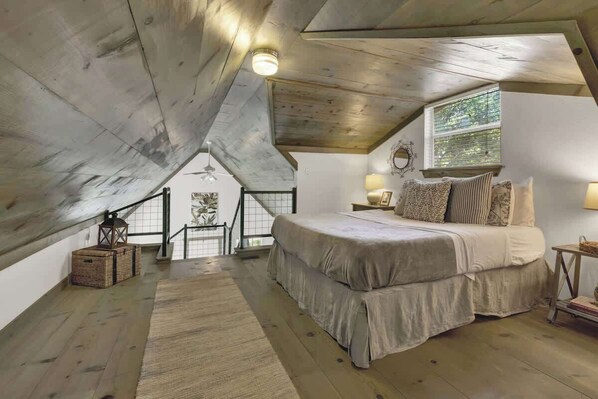 A bedroom with a sloped ceiling, a large bed, a window with blinds, and a staircase leading to an upper level.