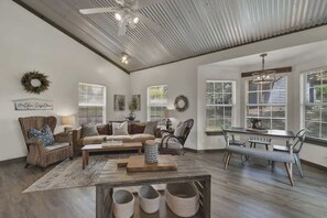 Ein großzügiges Wohnzimmer mit hoher Decke, einem Essbereich mit Bank und Tisch sowie großen Fenstern mit Jalousien.