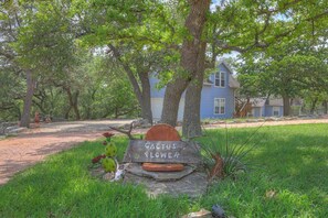 A rustic wooden sign with the text "Cactus Flower" surrounded by greenery and a house in the background. - Cactus Flower Lodge-Pet Friendly-Starlit Sky Views (Fredericksburg)