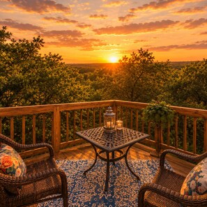 A wooden deck with wicker furniture and a table with candles, overlooking a forested landscape at sunset. - Cactus Flower Treehouse-Pet Friendly-Scenic Views! (Fredericksburg)