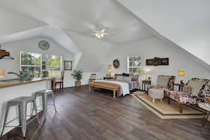 Ein geräumiges Schlafzimmer im Loft-Stil mit einem großen Bett, einem Schreibtisch und einem Sessel. Ein Fenster bietet Blick auf Bäume, ein Deckenventilator sorgt für Luftzirkulation und eine Uhr ist an der Wand angebracht.