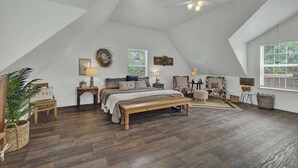 A spacious attic bedroom with a large bed, a wooden bench, a ceiling fan, and a view of the outdoors through a window. - Cactus Flower Treehouse-Pet Friendly-Scenic Views! (Fredericksburg)