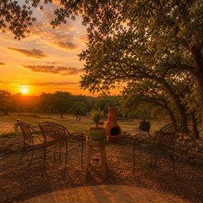 An outdoor seating area with chairs and potted plants, overlooking a natural landscape with trees and a sunset.