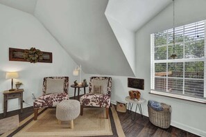 Ein Zimmer mit schrägem Dach, zwei Stühle mit Kuhmuster, ein kleiner runder Hocker, ein Beistelltisch mit Lampe, ein Korbwäscher und ein Fenster mit Jalousien.