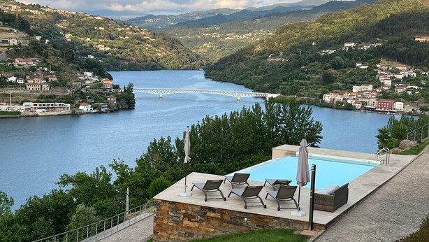 A pool area with lounge chairs and a stone wall, overlooking a river with a bridge and hills in the background.