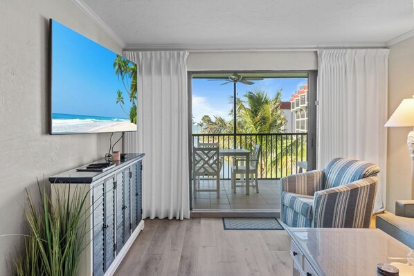 A modern living room with a flat-screen TV, a sofa, a dining table, and a balcony with a view of the ocean and palm trees.