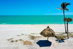 A beach with a thatched umbrella, palm trees, and a clear blue ocean.
