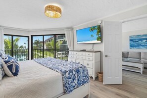 Un dormitorio con cama, cómoda, televisor y balcón con vista a palmeras y un edificio.