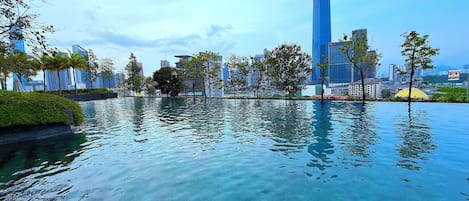A modern urban landscape with a large body of water reflecting buildings, surrounded by greenery and palm trees.