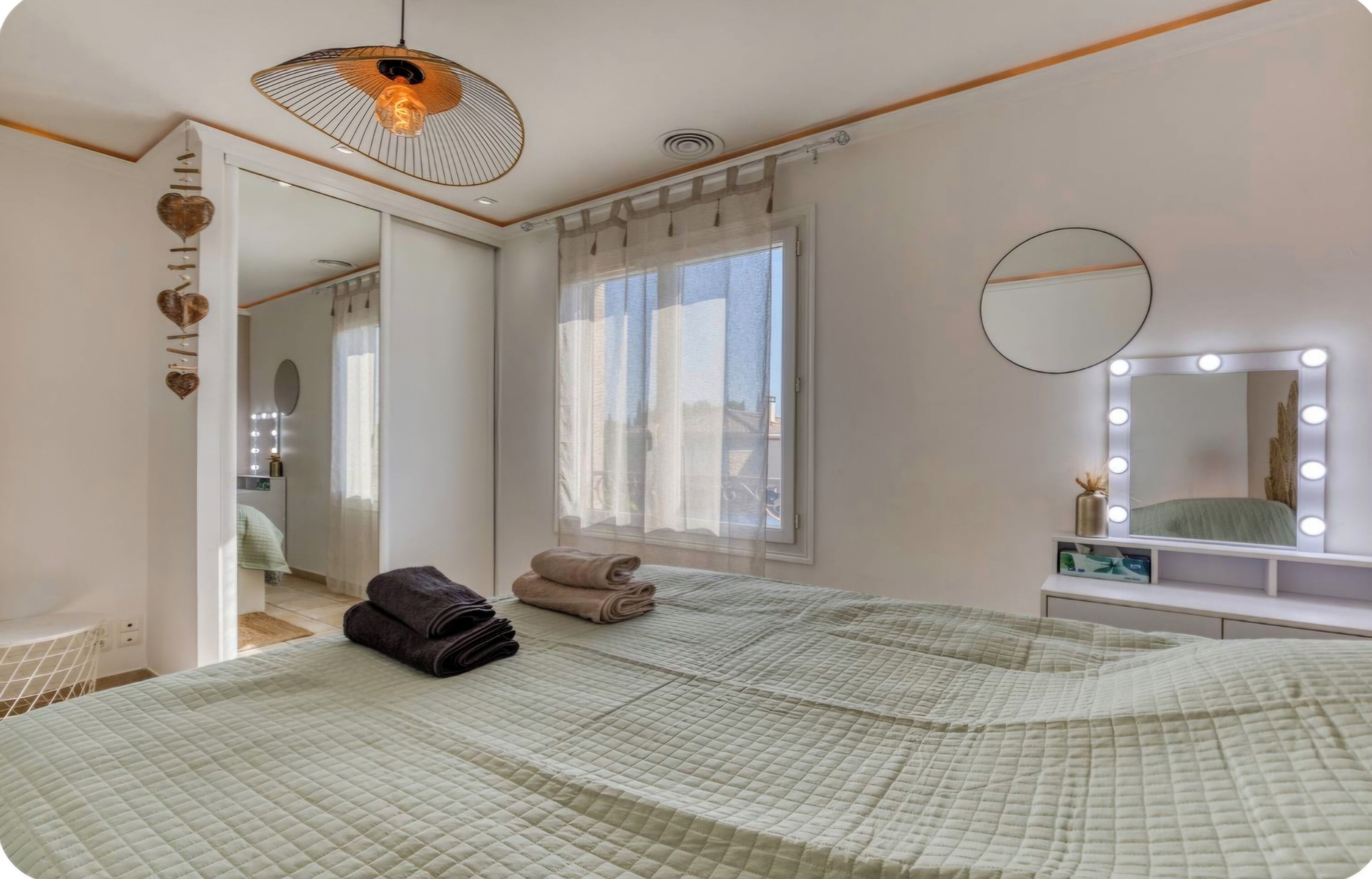 A bedroom with a large bed, a vanity with a mirror and lights, a window with curtains, and a hanging light fixture.